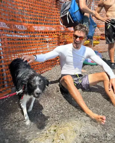 A smiling man in sunglasses and a white shirt sits on wet ground, petting a black and white dog on a leash—reminiscent of post-Amalfi caves kayak adventure relaxation. Behind them, two shirtless men stand near an orange safety fence on a sunny day.