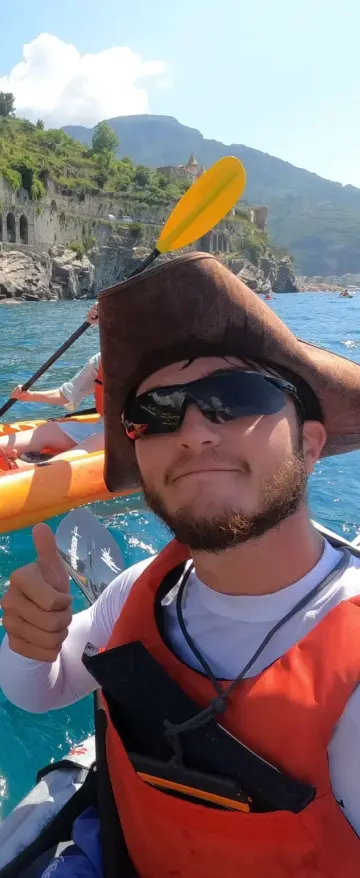 Two people in orange kayaks paddle on bright blue water near a rocky coastline with terraced hills on an Amalfi Coast Kayak Tour. One wears a pirate hat and gives a thumbs up, while the other smiles at the camera. Both wear life jackets.