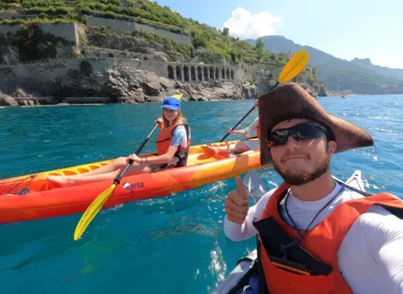 Two people in orange kayaks paddle on bright blue water near a rocky coastline with terraced hills on an Amalfi Coast Kayak Tour. One wears a pirate hat and gives a thumbs up, while the other smiles at the camera. Both wear life jackets.
