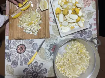 A person chops lemons on a wooden board near a basket of whole lemons, a tray with lemon peels, and a bowl of chopped lemon pieces—all set on a floral tablecloth reminiscent of picnics after the best hikes Amalfi Coast offers.