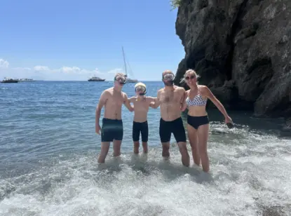 Four people wearing swimsuits and snorkeling masks stand together in shallow ocean water near a rocky cliff, as boats from the Best Kayak Tours Amalfi and a sailboat are visible in the sunny background.