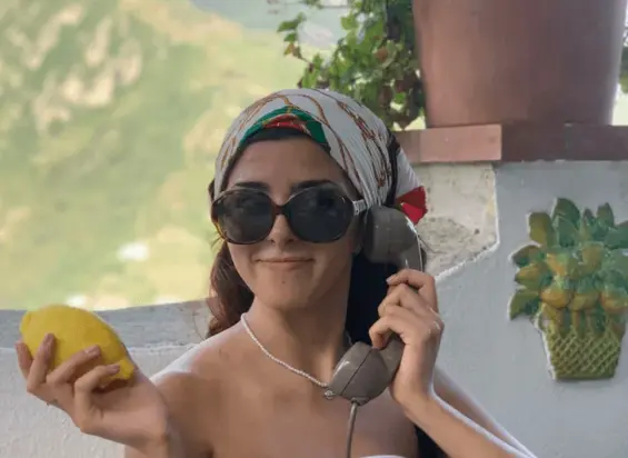 Una donna con occhiali da sole e un foulard colorato tiene in una mano un limone e nell'altra una cornetta telefonica vecchio stile, catturando lo spirito vibrante di una camminata tra Maiori e Minori mentre si trova all'aperto accanto a un muro bianco con una decorazione di ananas.