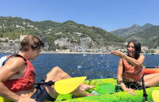 A woman splashes water playfully at a man as they enjoy coastal exploration by kayak on a blue lake, with another woman paddling behind them and green hills and mountains under a clear sky. A woman splashes water playfully at a man as they enjoy coastal exploration by kayak on a blue lake, with another woman paddling behind them and green hills and mountains under a clear sky.