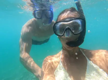 Two people wearing snorkel masks and swimwear take an underwater selfie while snorkeling in crystal clear water, with sunlight creating patterns on their skin.