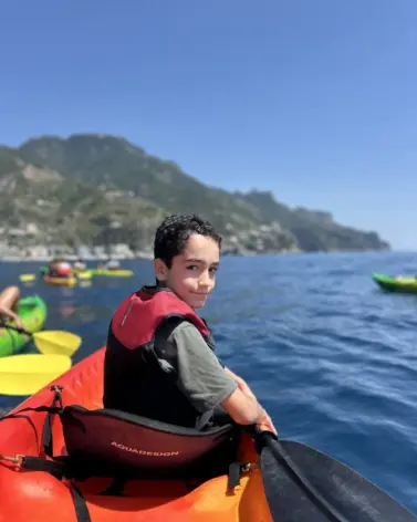 A young boy in a red life jacket paddles a kayak on calm blue water during a guided kayak Amalfi tour, with other kayaks and green mountains in the background under a clear sky.