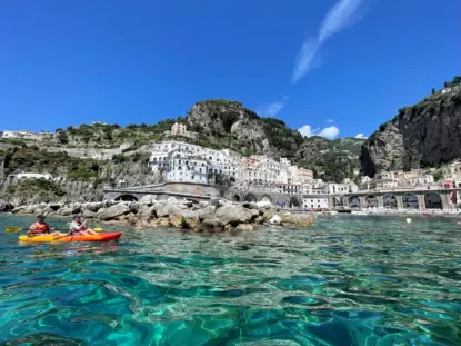 Two people enjoy a guided sea kayak adventure on clear turquoise water near a rocky shore, with white hillside buildings and lush greenery under a bright blue sky in a stunning Amalfi coastal town.