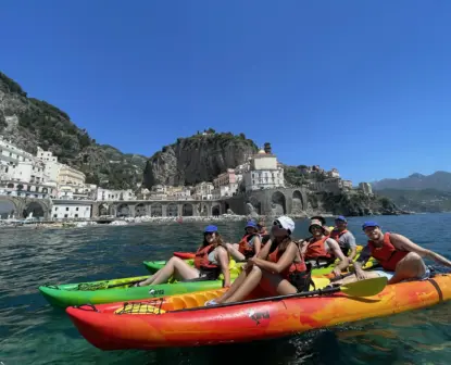 A group of six people in colorful kayaks on clear blue water, enjoying a kayak rental Amalfi Coast adventure near a coastal town with historic buildings, arches, and steep hills under a bright blue sky.