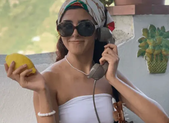 A woman wearing a white tube top, sunglasses, and a colorful headscarf channels the charm of a lemon groves walking tour in Maiori as she holds a lemon and an old-fashioned phone near a white wall with pineapple decor.