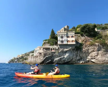 Two people in a red and yellow kayak paddle on clear blue water along the Amalfi Coast near rocky cliffs with historic buildings and greenery, under a bright blue sky, possibly spotting vibrant marine life below.