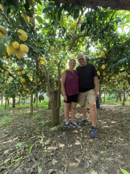 A smiling couple stands together under lush lemon trees filled with ripe yellow lemons along the scenic Path of the Lemons Amalfi, sunlight filtering through the leaves in a picturesque orchard.