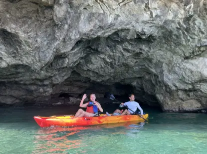Two people in life jackets paddle a red and yellow kayak near the entrance of a rocky cave, surrounded by clear, turquoise water on a Sea Kayaking Amalfi Coast adventure. One waves at the camera while the other holds a paddle.
