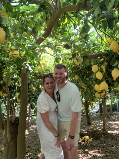 A smiling couple poses together under a canopy of lemon trees along the Sentiero dei Limoni hike. Sunlight filters through the leaves as they enjoy the trail; she wears a white dress and sunglasses, and he’s in a white t-shirt and shorts.