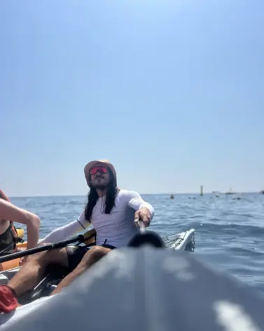 A man wearing sunglasses and a hat paddles a kayak on calm blue water under a clear sky during a snorkeling experience in Amalfi. The photo is taken from the front of the kayak, with another person partially visible on the left.
