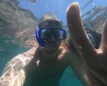 A person snorkeling our Amalfi Coast wears a snorkel mask underwater, making a peace sign while taking a selfie; sunlight shimmers on their face and the vibrant blue water around them.