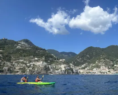 Two people paddle a green kayak in bright blue water, with green hills, traditional lemon terraces of Amalfi, and a coastal town in the background under a blue sky with a few fluffy white clouds.